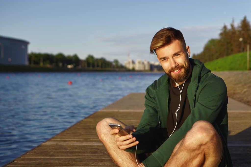 Smilende ung mand sidder ved floden og lytter til musik med høretelefoner og en smartphone