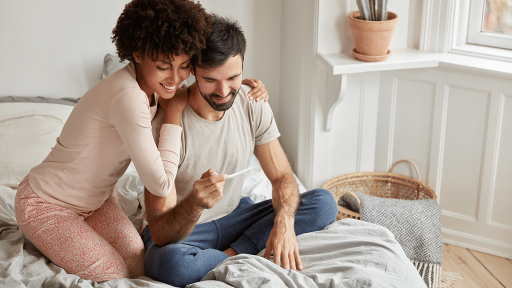 A happy couple sitting on a bed, smiling and holding a pregnancy test in a bright and cozy home setting.