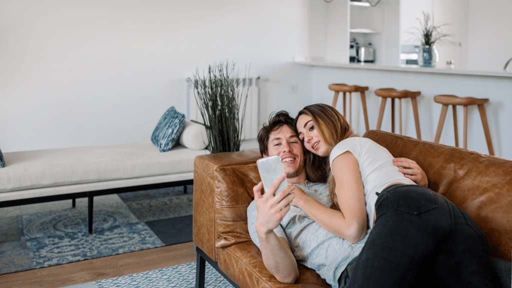 A couple lounging on a couch, smiling while looking at a smartphone, symbolizing the ease and comfort of at-home male fertility testing.