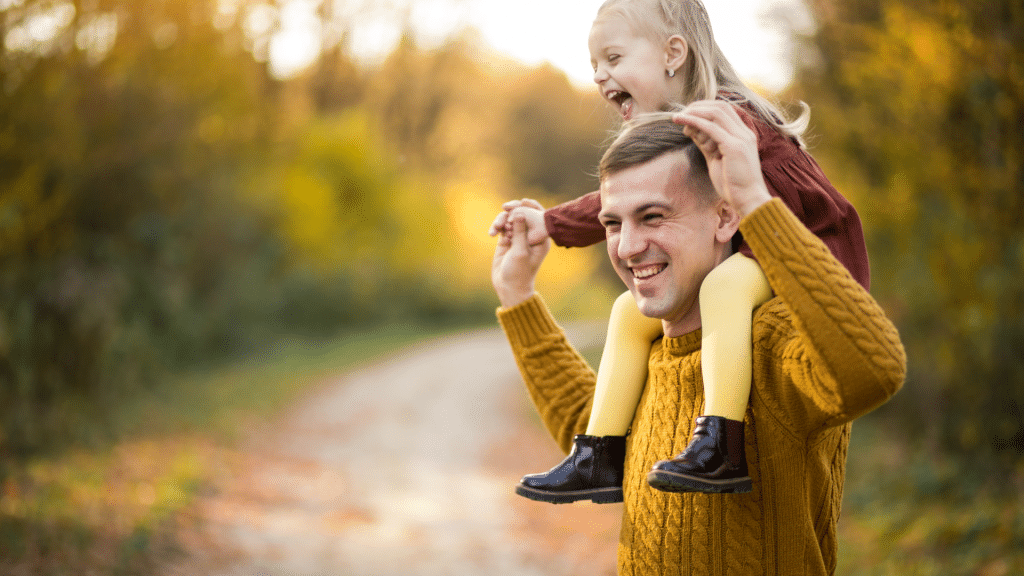 Padre sonriente que lleva a hombros a su alegre hija durante un paseo por la naturaleza, representando la vitalidad, la longevidad y la salud familiar.