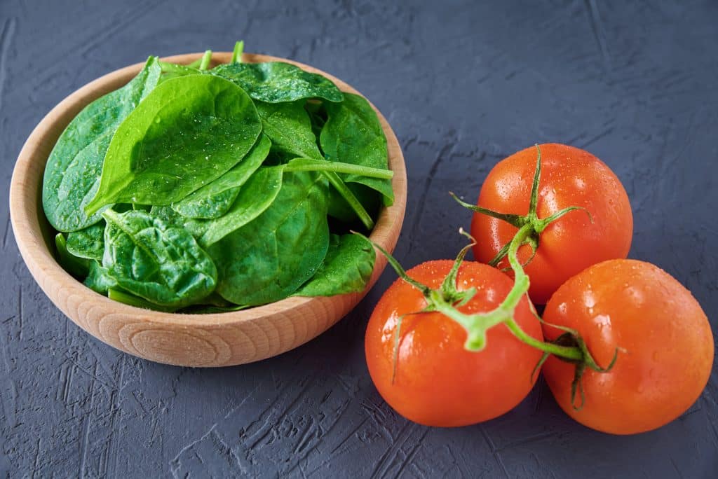 Hojas de espinacas frescas en un cuenco de madera con tomates enteros sobre fondo oscuro, ricas en folato y licopeno para la salud reproductiva masculina.