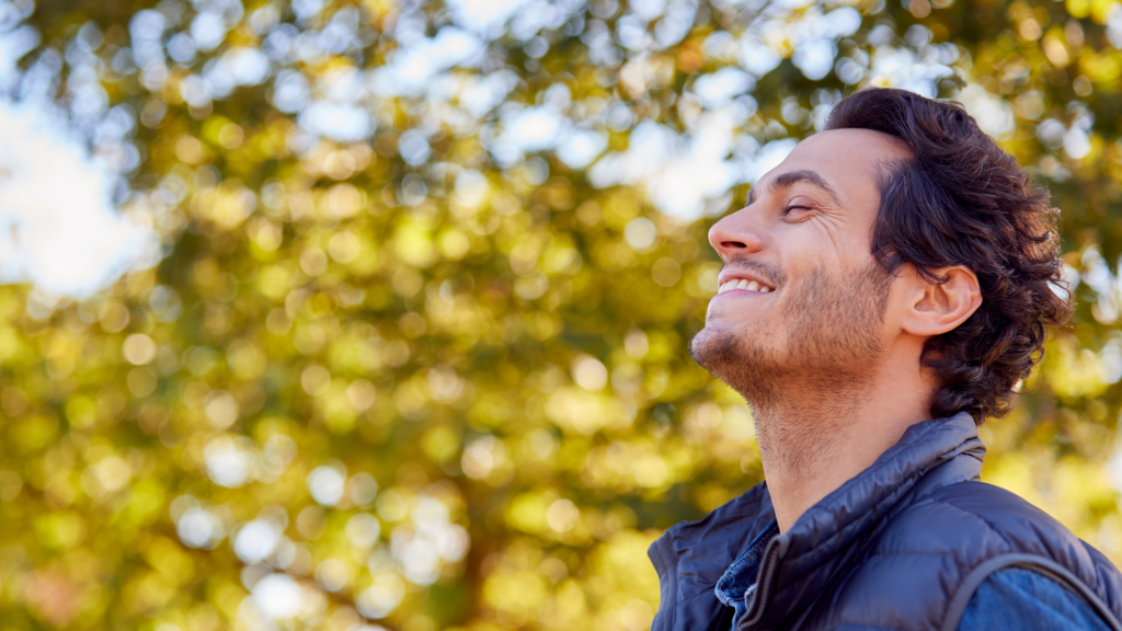 Semana de Pentecostés - Hombre relajado sonriendo con los ojos cerrados en un parque verde, optimizando su fertilidad primaveral durante un descanso vacacional.
